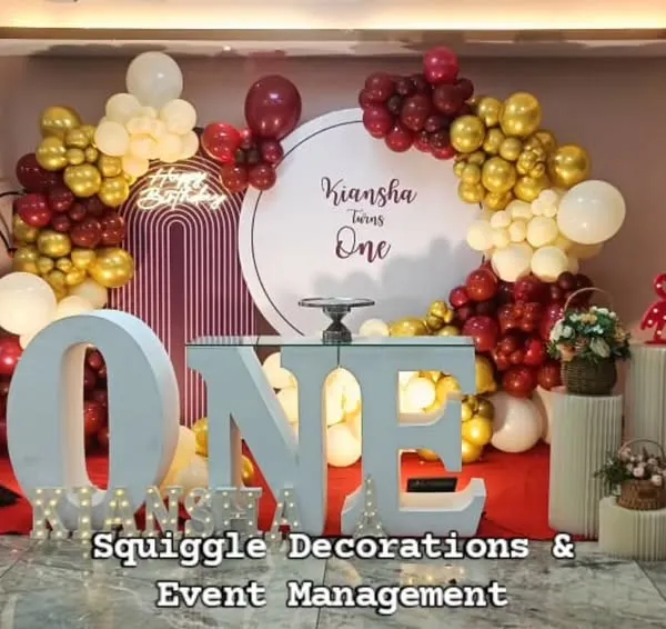 A sweet first birthday setup with a large marquee 'ONE', a custom circular backdrop, and a red and gold balloon garland.
