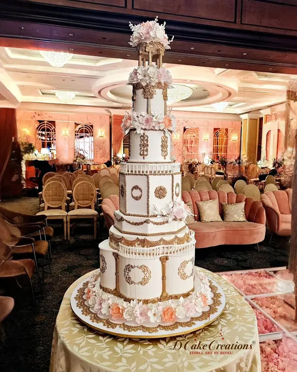 A full view of our architectural engagement cake, standing tall in a beautifully decorated ballroom. The design features multiple tiers with distinct shapes, ornate gold detailing, and a floral gazebo topper, creating an unforgettable presence.