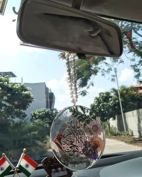 This custom car hanging with Ayat ul Kursi is shown in its natural setting, hanging from a rearview mirror. It includes real flowers and glitter, catching the sunlight beautifully.