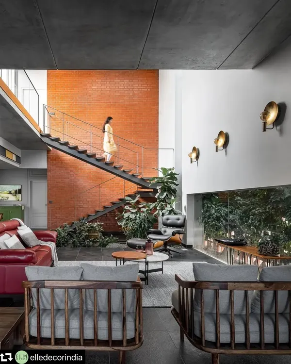 A sleek metal staircase ascends alongside a warm, exposed brick wall in the double-height living area. This feature connects the home's two levels while acting as a backdrop to the indoor courtyard, blending industrial and natural elements.