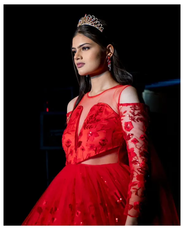A princess-style red gown captured in its full glory on the runway. The lighting highlights the sheer, embroidered sleeves and the sparkle of the tiara, creating a fairy-tale moment during the fashion show.