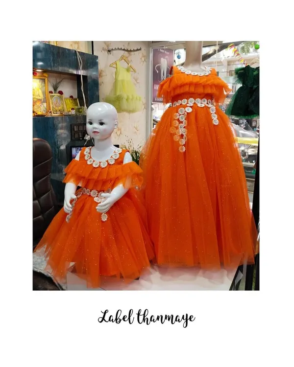 A mother-daughter twinning set in bright orange tulle, displayed on mannequins. Both the gown and the frock feature an off-shoulder neckline with white floral applique details.