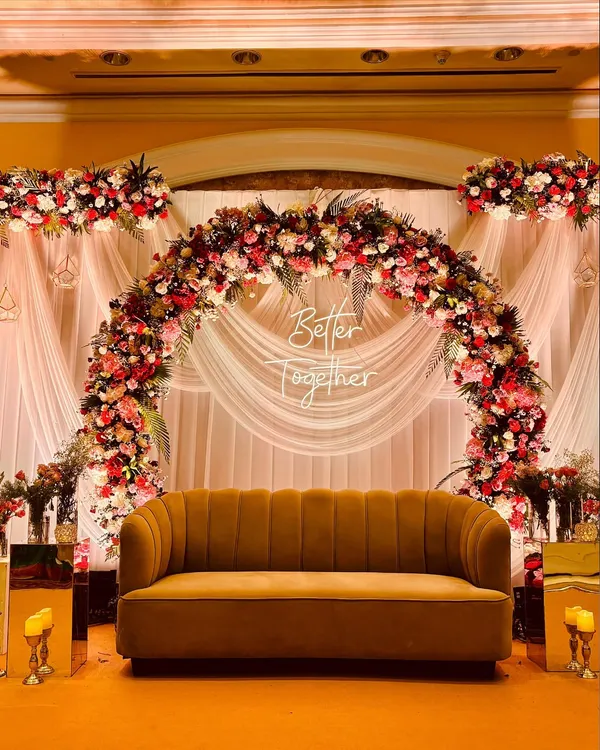 A romantic stage backdrop featuring a lush floral arch of red and pink roses, with soft white drapes and a "Better Together" sign, perfect for couple's photos.