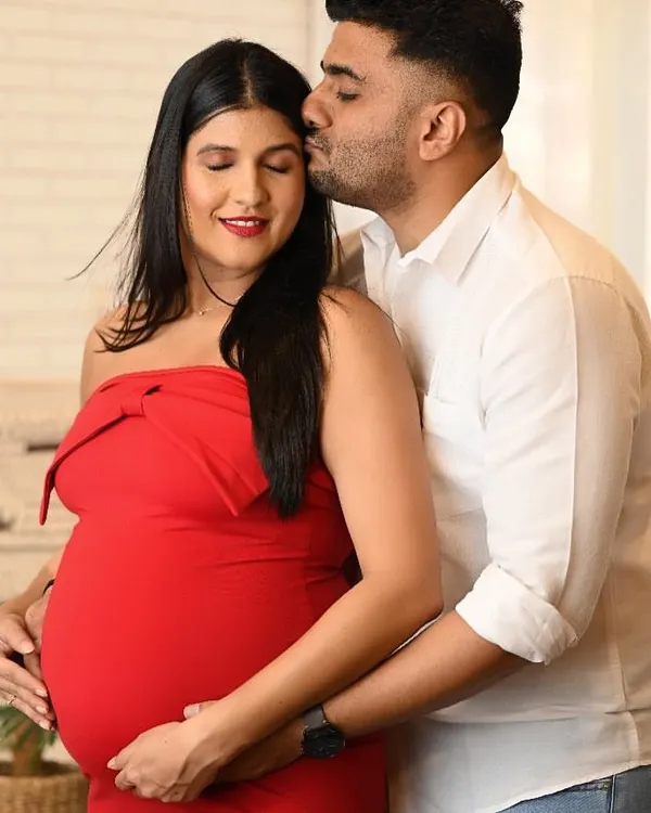 A tender moment between an expecting couple, with the partner kissing the mother-to-be's forehead. The woman is wearing a vibrant red dress, adding a pop of color to this studio maternity portrait.