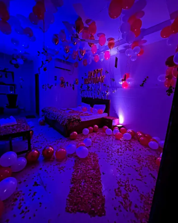 A full room decorated for a birthday surprise, with red and white balloons, a rose petal pathway, and moody blue lighting.