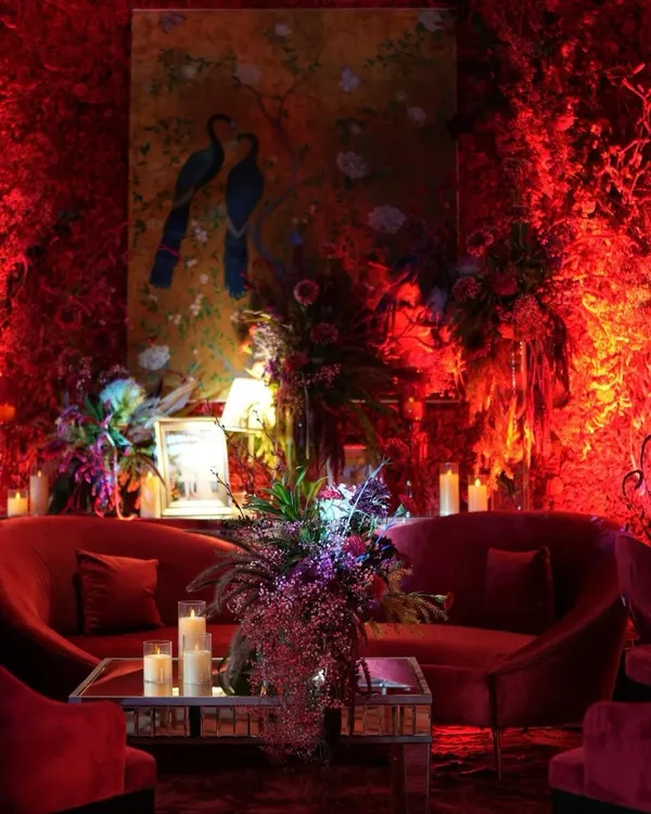 A luxurious lounge area drenched in shades of red. The space features plush red velvet sofas, a mirrored coffee table, and walls covered in red foliage, all illuminated by warm, dramatic lighting. A hand-painted peacock artwork adds a touch of artistry to the passionate design.