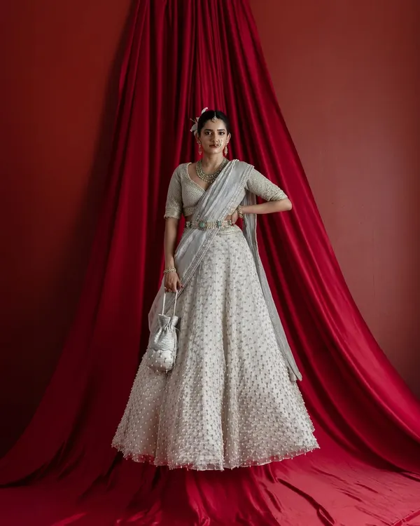 A full-length view of the magnificent ivory bridal lehenga, complete with a matching potli bag. The entire ensemble, from the detailed skirt to the draped dupatta, is designed to create a cohesive and breathtaking bridal statement against a dramatic red backdrop.