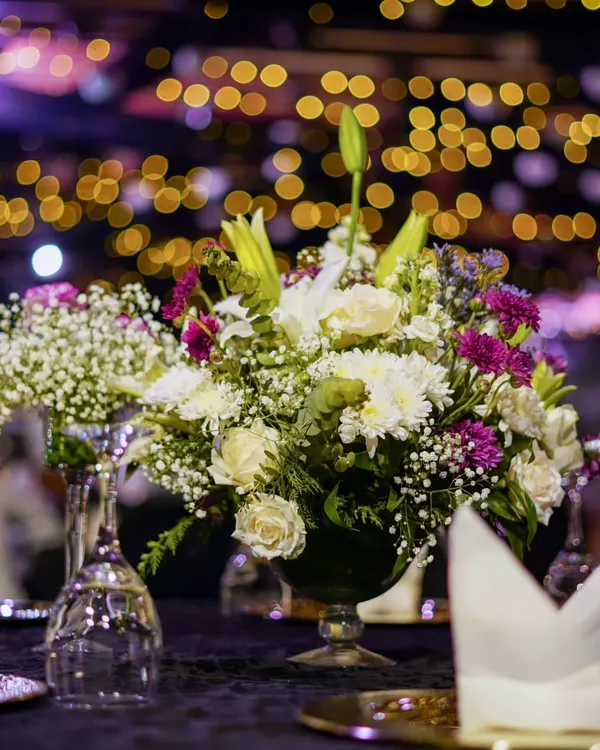 A close-up of a lush floral centerpiece, with a beautiful bokeh effect from the event lighting in the background. We use a mix of flowers like lilies, roses, and chrysanthemums to create texture and depth in our arrangements.