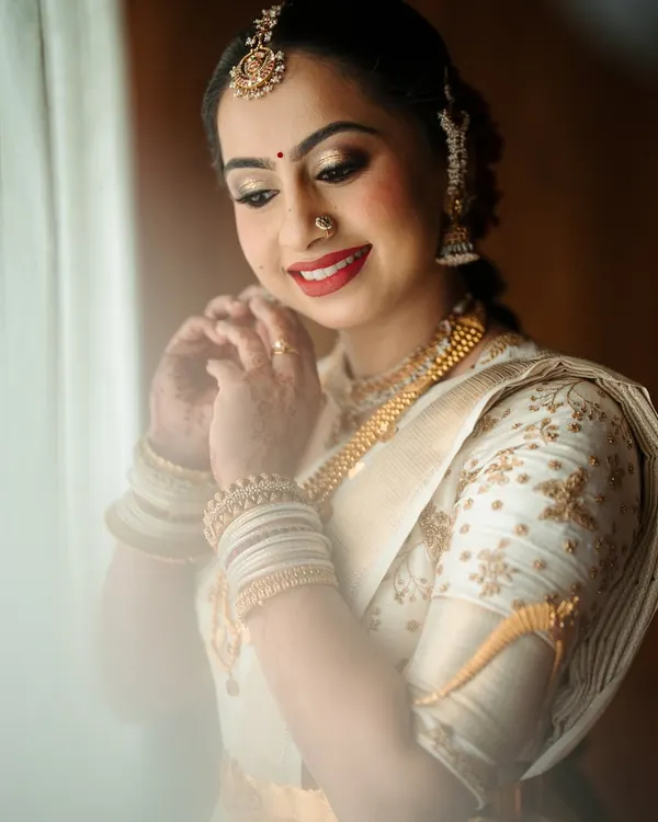 A beautiful bride getting ready in an elegant off-white Kanchipuram saree. Her custom blouse features delicate gold floral embroidery that perfectly matches her traditional gold jewelry.