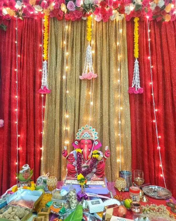 A vibrant home Ganpati mandap featuring a bright red idol. The backdrop combines a shimmery golden curtain with rich red drapes, accented by fairy lights and hanging floral tassels for a classic festive look.
