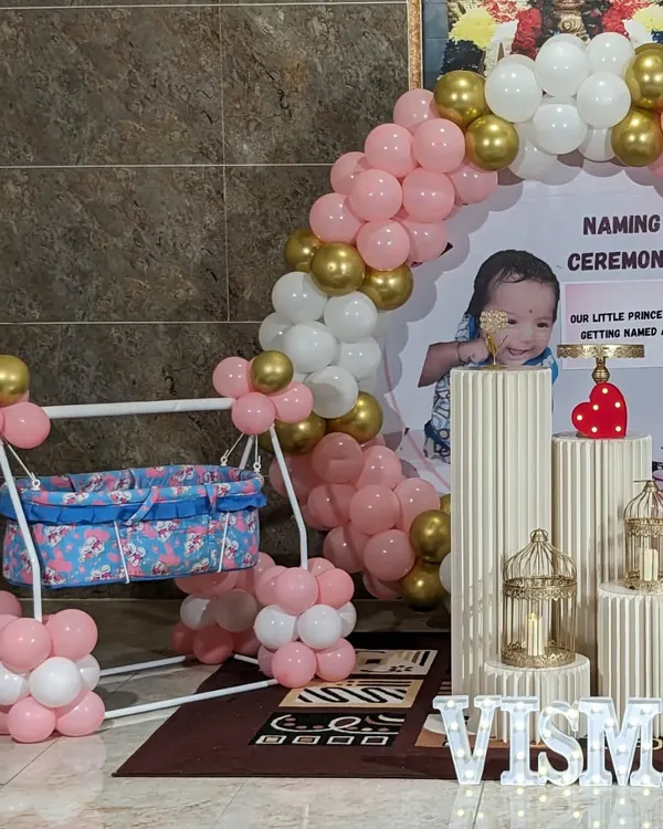 A modern naming ceremony setup featuring a personalized backdrop with the baby's photo. The decor includes a pink, white, and gold balloon arch, a decorated cradle, and illuminated letters spelling the name.