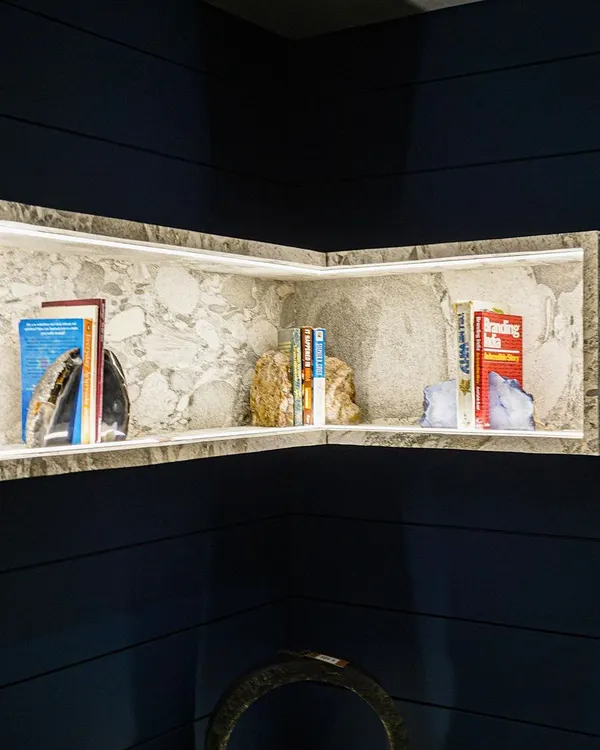 This custom corner bookshelf is crafted from a single piece of stone, with integrated LED lighting that casts a warm glow on the displayed books and objects. Set against a dark blue grooved wall, it stands out as a piece of functional sculpture.