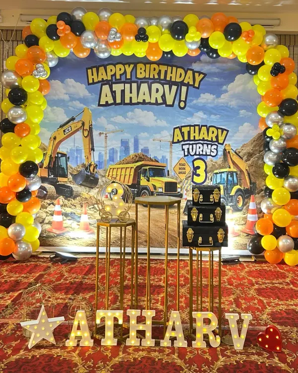 A full-scale construction-themed 3rd birthday for Atharv. This setup includes a large, detailed backdrop of a construction site, a balloon arch in yellow, black, and orange, and marquee letters.