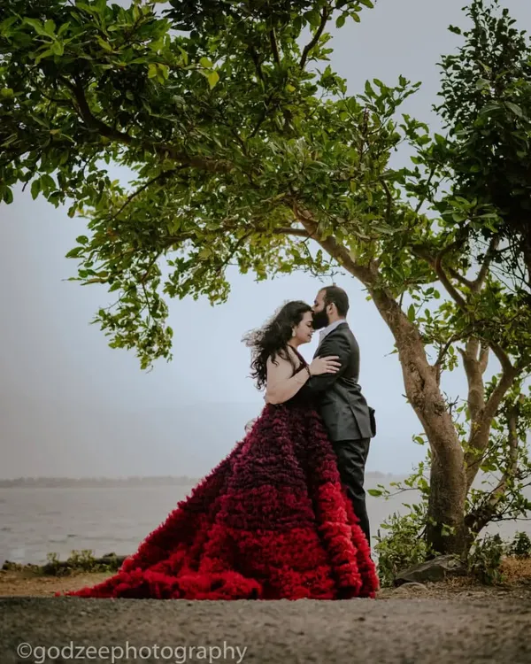 A truly romantic pre-wedding moment captured under a lush green tree by the water. The wind catching her hair and the stunning red ombre gown makes this shot feel like a movie scene.