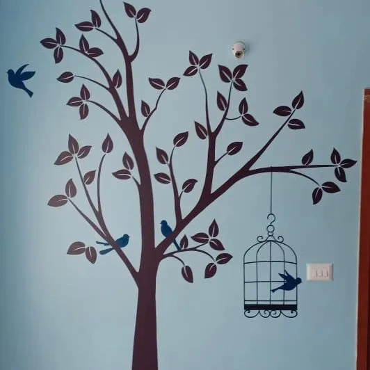 A large tree stencil in dark brown on a light blue wall creates a serene nature scene. The design includes small birds perched on branches and a decorative birdcage, adding a whimsical touch.