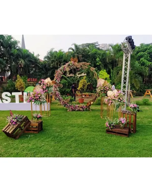 An open-ground wedding setup featuring a rustic-chic theme. The centerpiece is a large circular backdrop with the words 'Better Together', surrounded by floral arrangements on wooden crates and stands.