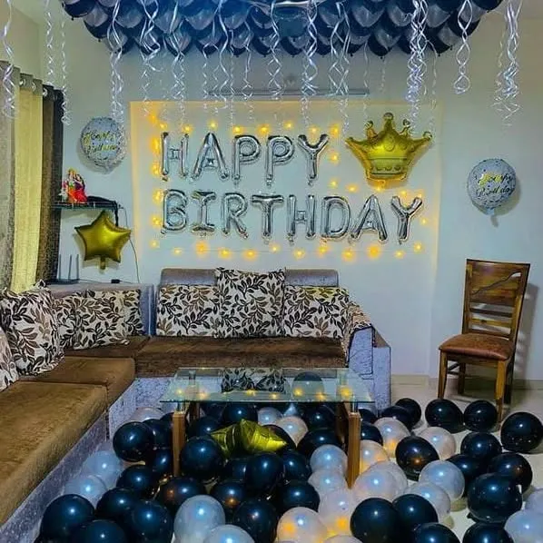 A classic living room birthday setup. This decoration includes 'Happy Birthday' foil letters with a crown, a fairy light backdrop, and black and silver balloons on the ceiling and floor.