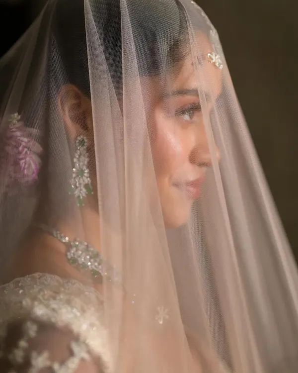 An ethereal bridal look behind a delicate veil. Dressed in a dreamy white and pink lehenga with emerald diamond jewels, the makeup is subtle, clean, and natural to enhance her glow.
