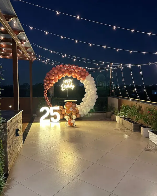 A stunning rooftop terrace party for a 25th birthday, featuring a rose gold and white balloon ring under string lights against the night sky.