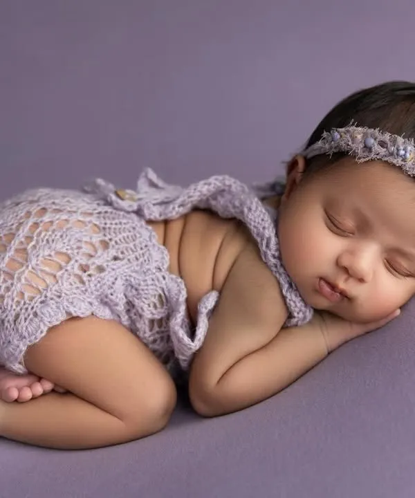 A classic side lying pose on a soft lavender backdrop. The baby is dressed in a delicate lace romper and a matching headband, creating a sweet and serene portrait.