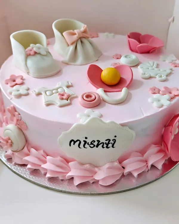 A sweet welcome cake for a baby girl, decorated with edible baby shoes, a pacifier, and other cute baby items.