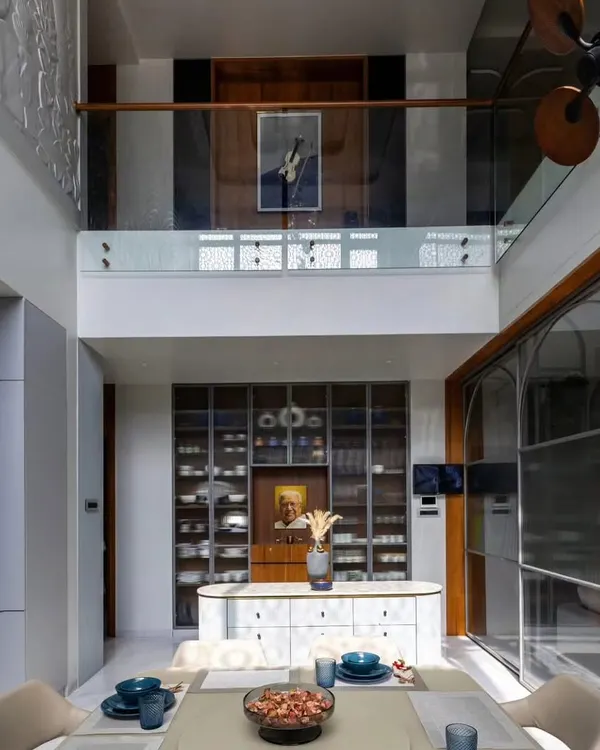 A grand dining area in a duplex home, featuring a double-height ceiling and a glass-paneled mezzanine. The custom crockery unit and island counter add both functionality and a touch of luxury.