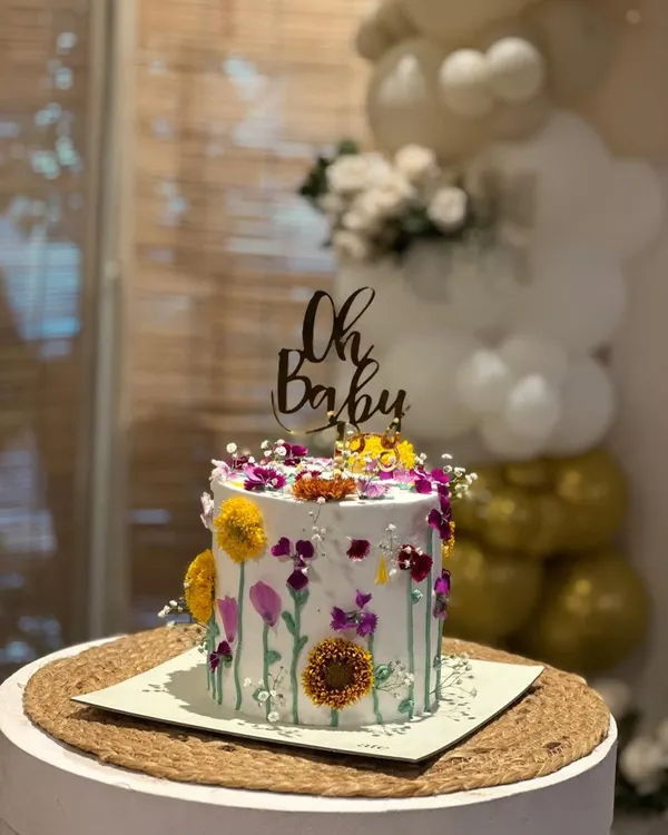 A simple and elegant "Oh Baby" topper in dark wood or acrylic, sitting atop a beautiful wildflower cake. It's the perfect touch for a rustic or boho-themed baby shower.
