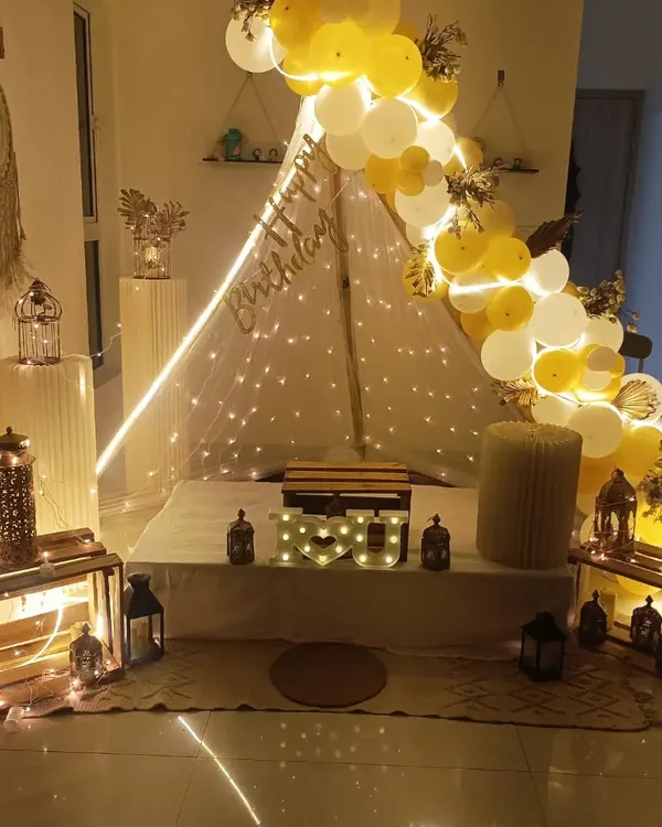 A beautiful and bright birthday cabana setup in white and yellow. The tent is surrounded by decorative lanterns, wooden crates, and a lovely balloon arch.
