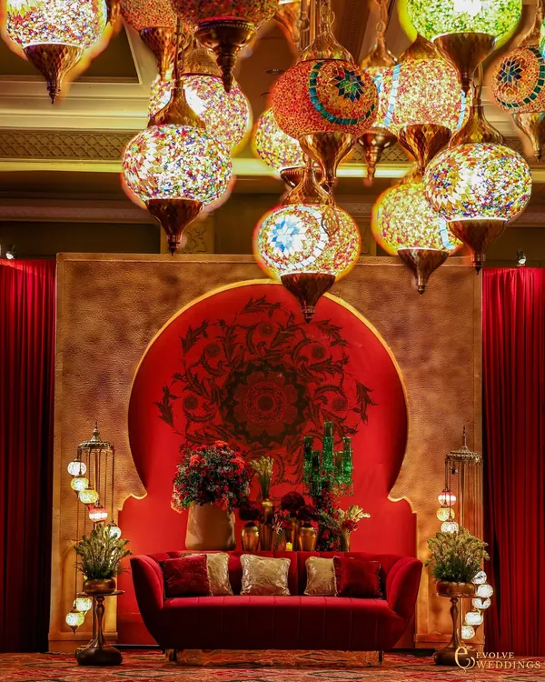 A central seating area designed for a Sufi-themed night. A plush red velvet sofa is set against a circular mandala backdrop, while a cascade of colorful Turkish mosaic lamps provides an intimate and magical glow.