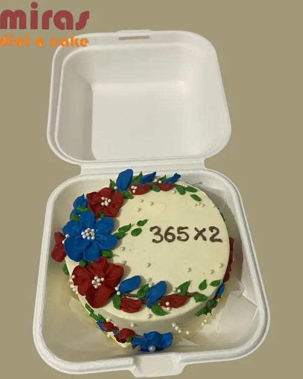 Celebrating a second anniversary, this bento cake features a wreath of delicate, hand-piped flowers in red and blue. It's a simple yet elegant way to mark a special occasion like "365 x 2" days together.