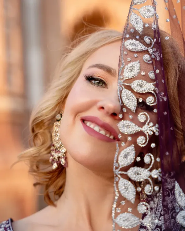 A joyful close-up portrait of a model in a traditional Indian lehenga. She peeks from behind an intricately embroidered dupatta, with a genuine smile that lights up the frame.