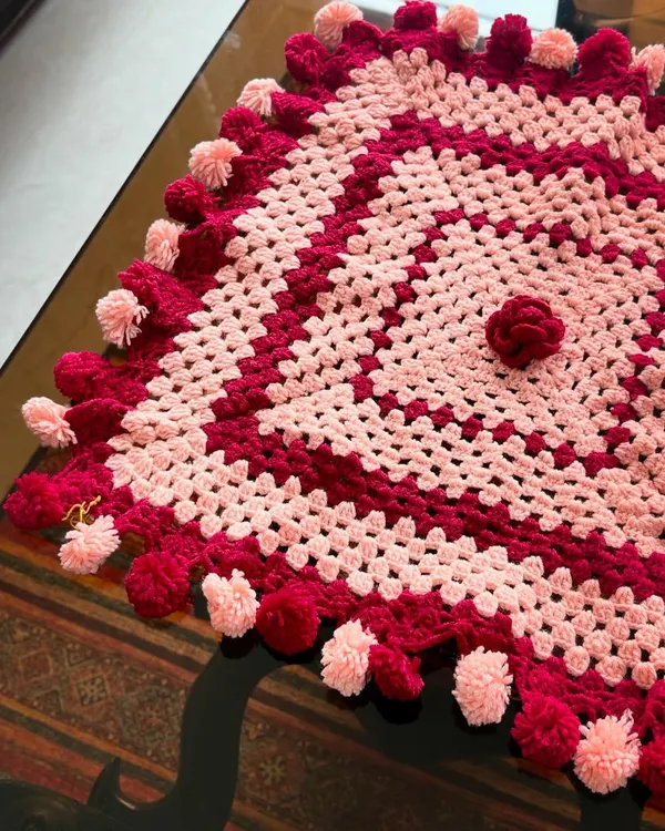 This square tablecloth in shades of pink and red is finished with playful pompoms, making it a festive addition to any table setting.