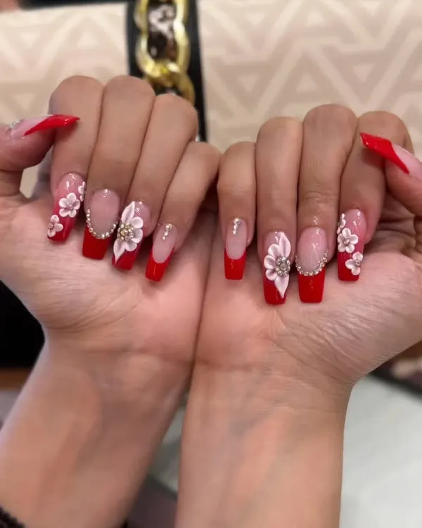 A bold and festive look featuring bright red gel extensions with a modern French tip style, accented with intricate, hand-crafted 3D white flowers and gold details.