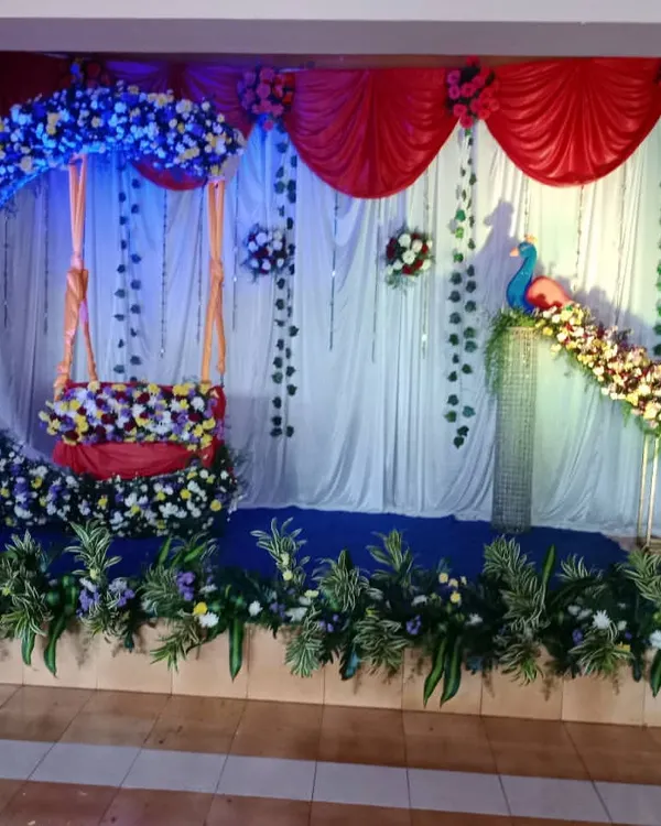 A full stage setup for a naming ceremony with a floral crescent moon swing and a peacock figure. The design uses a mix of blue, white, and yellow flowers with hanging green vines.