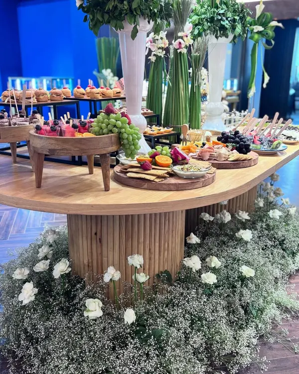 A breathtaking grazing table, designed as the centerpiece for a luxury event. This opulent display features multiple tiers of artisanal cheeses, fresh fruits, and gourmet bites, surrounded by a bed of fresh baby's breath and white roses, embodying pure elegance and abundance.
