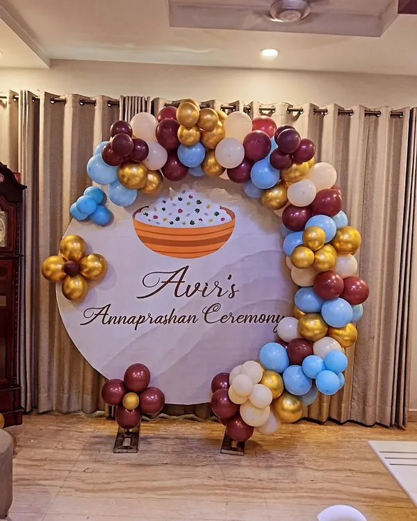 A personalized backdrop for "Avir's Annaprashan Ceremony." This design features a circular backdrop with a rice bowl graphic, framed by a balloon arch in a unique color combination of maroon, blue, and gold.