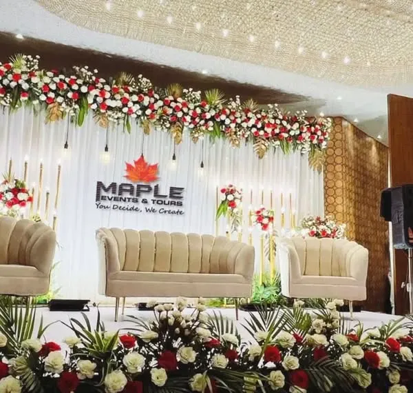 A beautiful stage setup for a formal event, featuring plush seating and a backdrop adorned with red and white floral arrangements. The combination of hanging lights and elegant furniture creates a warm and inviting atmosphere.