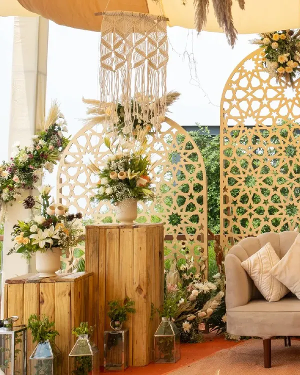 A perfect corner in our rustic-themed decor. This setup features a modern sofa, wooden block tables, and beautiful floral arrangements, all framed by intricate jali-work panels and macrame hangings.