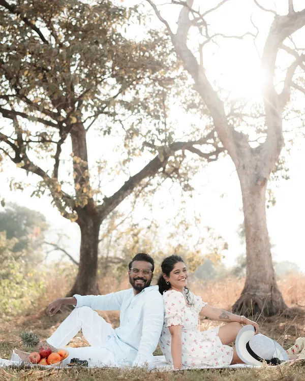 A couple sits back-to-back on a picnic blanket under a beautiful tree, smiling as the sun shines behind them. This shot captures the relaxed and joyful vibe of an outdoor pre-wedding session.