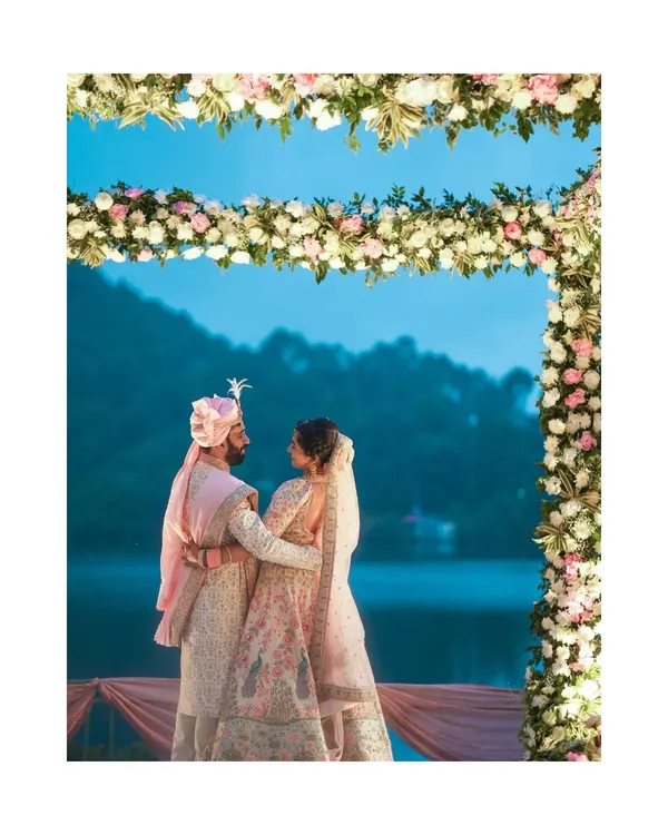 A stunning destination wedding portrait of a couple under a floral arch, with a serene lake and mountains at dusk. This is the magic of a Nainital wedding.
