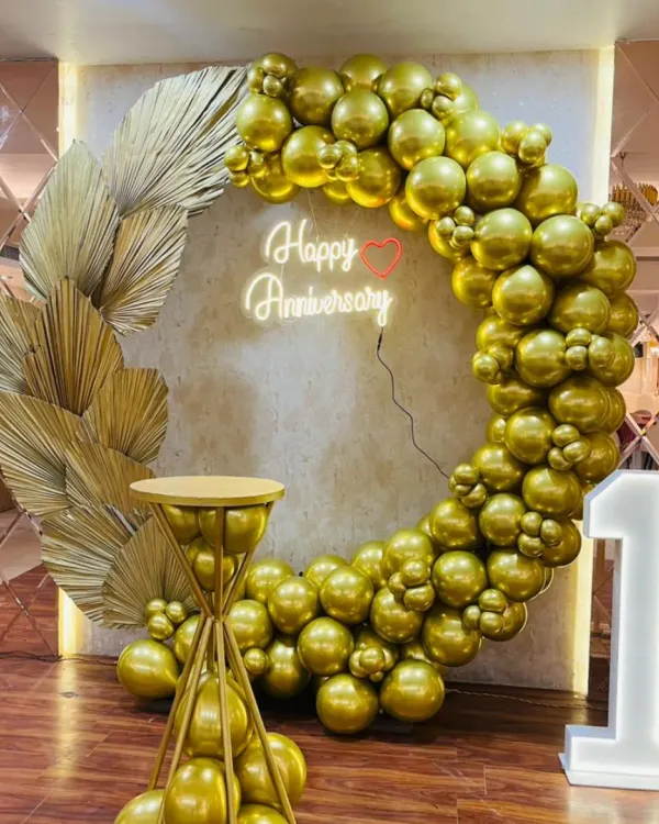 An all-gold theme for a first anniversary, featuring a circular balloon arch made of chrome gold balloons, a neon sign, and a matching cake pedestal. This setup, shown here for a first milestone, can be adapted for any anniversary year.