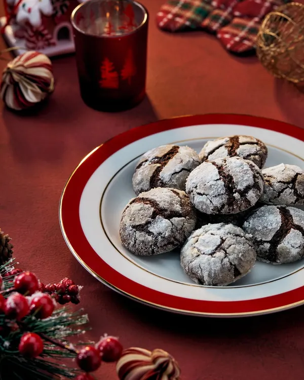 A cozy Christmas scene featuring chocolate crinkle cookies for Marriott Bonvoy. The warm lighting from the candle and the rich red tones of the plate and decor create a classic, festive atmosphere.
