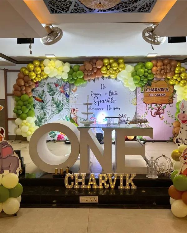 A grand jungle safari themed first birthday stage. The setup includes a large "ONE" prop, a backdrop with tropical leaves, and a balloon arch in earthy tones of green, brown, and yellow.