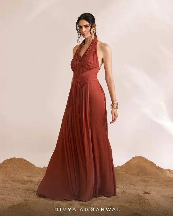 The Luna Dress in a rich rust color, designed with a graceful halter neck and an intricately pleated bodice. The flowing A-line skirt creates a silhouette of timeless elegance.