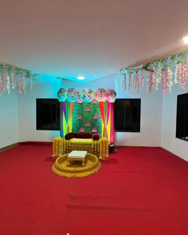 A wide view of a Haldi decoration setup we did in a hall. The space features a colorful stage backdrop, traditional seating, and hanging floral decorations from the ceiling.