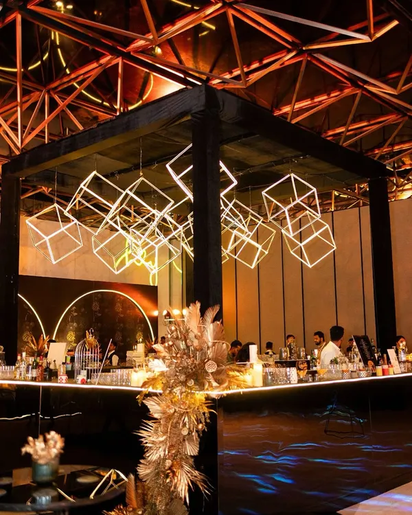 A modern, industrial-themed sangeet featuring a spectacular installation of glowing geometric cubes suspended over the main bar. The black and gold palette adds a layer of sophisticated glamour.