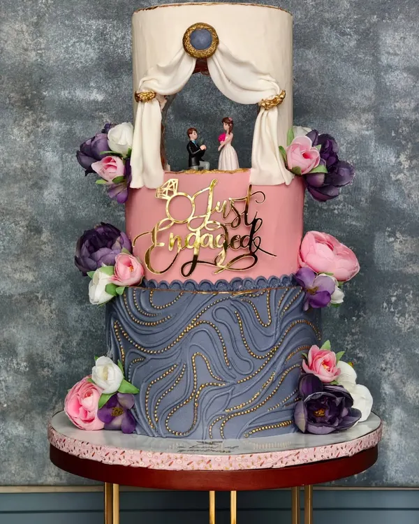 A full shot of the grand three-tiered engagement cake. The design combines a textured grey bottom tier, a smooth pink middle tier with a "Just Engaged" topper, and a top tier featuring a romantic diorama.