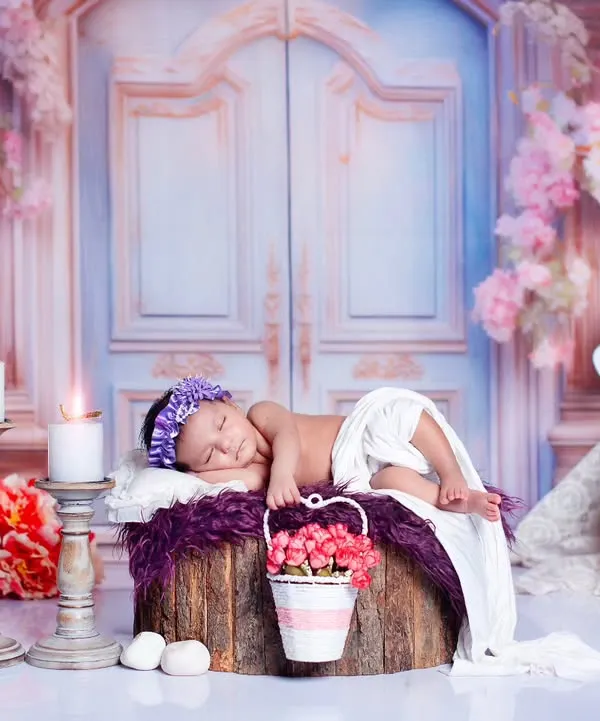 A newborn girl sleeps peacefully on a log, surrounded by a magical floral gate setup. The scene includes a unicorn figure and soft candlelight, creating a fairytale feel.