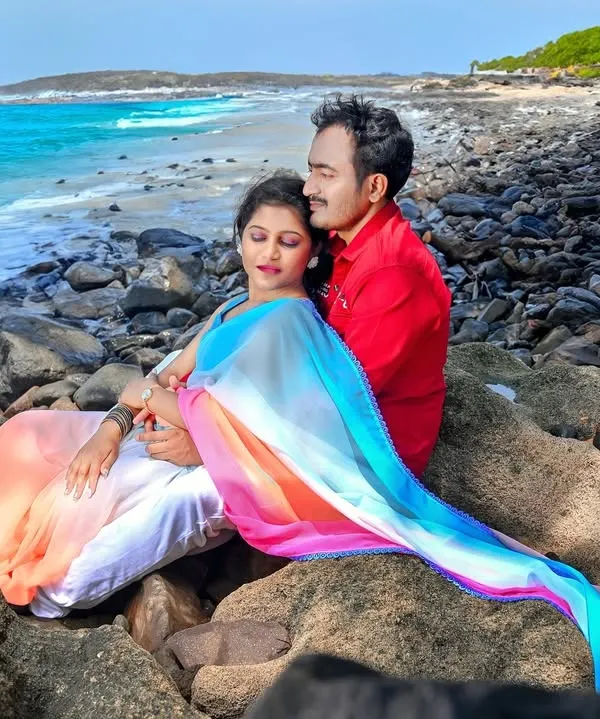 A serene pre-wedding portrait of a couple resting on coastal rocks. The vibrant, multi-colored dupatta flows in the breeze against the backdrop of the blue sea, creating a picturesque and romantic scene.