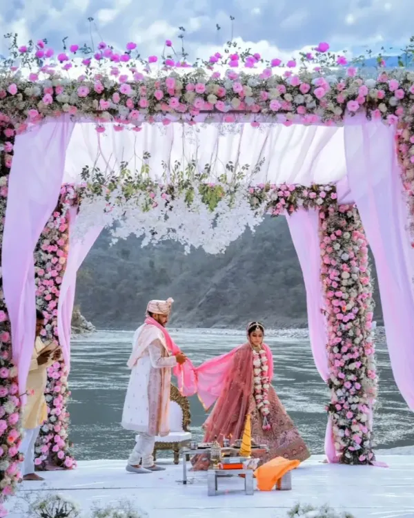 A beautiful moment from a riverside wedding in Rishikesh, with the couple under a mandap lavishly decorated with pink and white flowers. The natural landscape of the river and hills provides a truly unique setting.